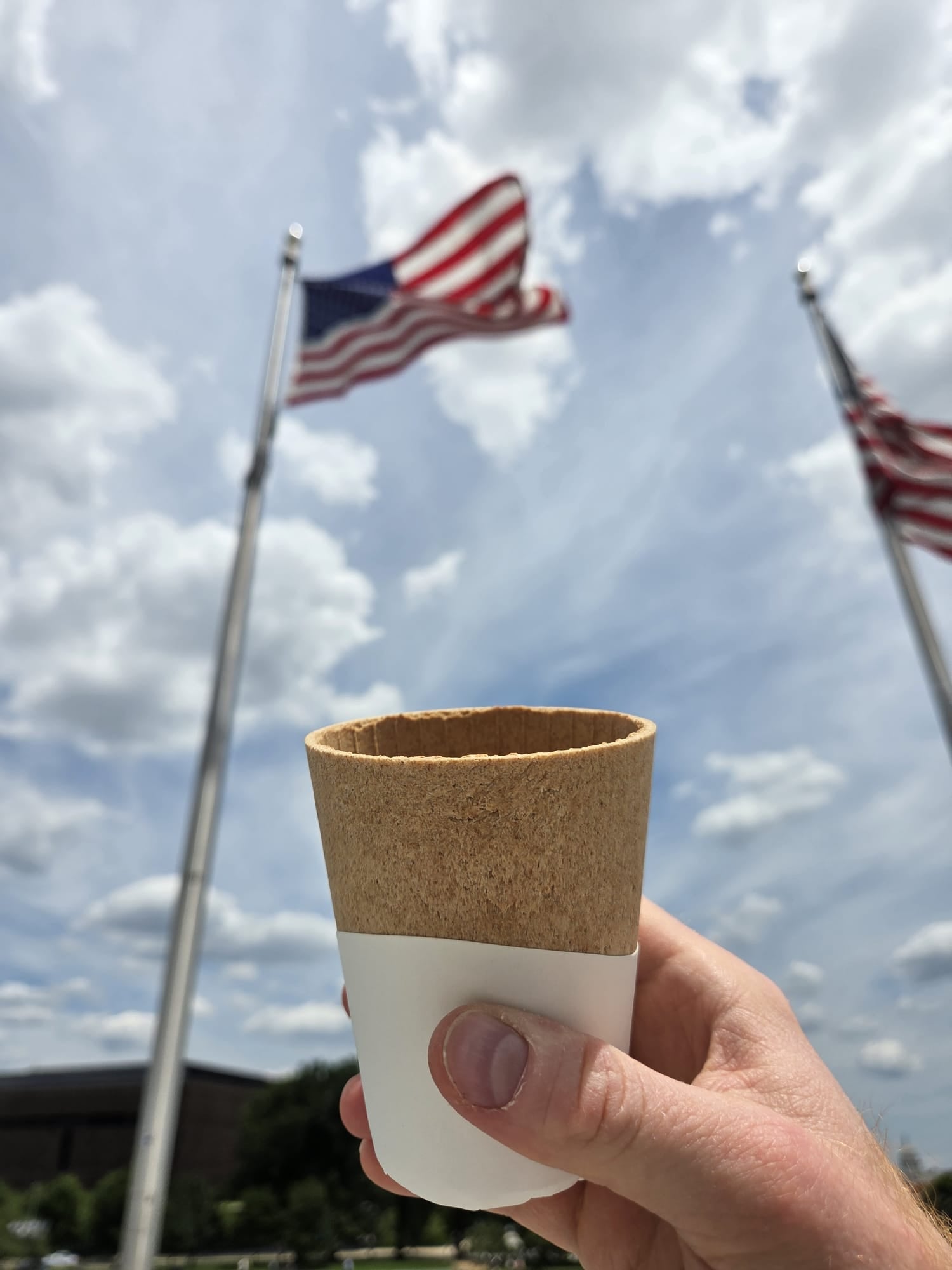 Hand holding a paper cup with a cork sleeve against a backdrop of American flags and a blue sky.