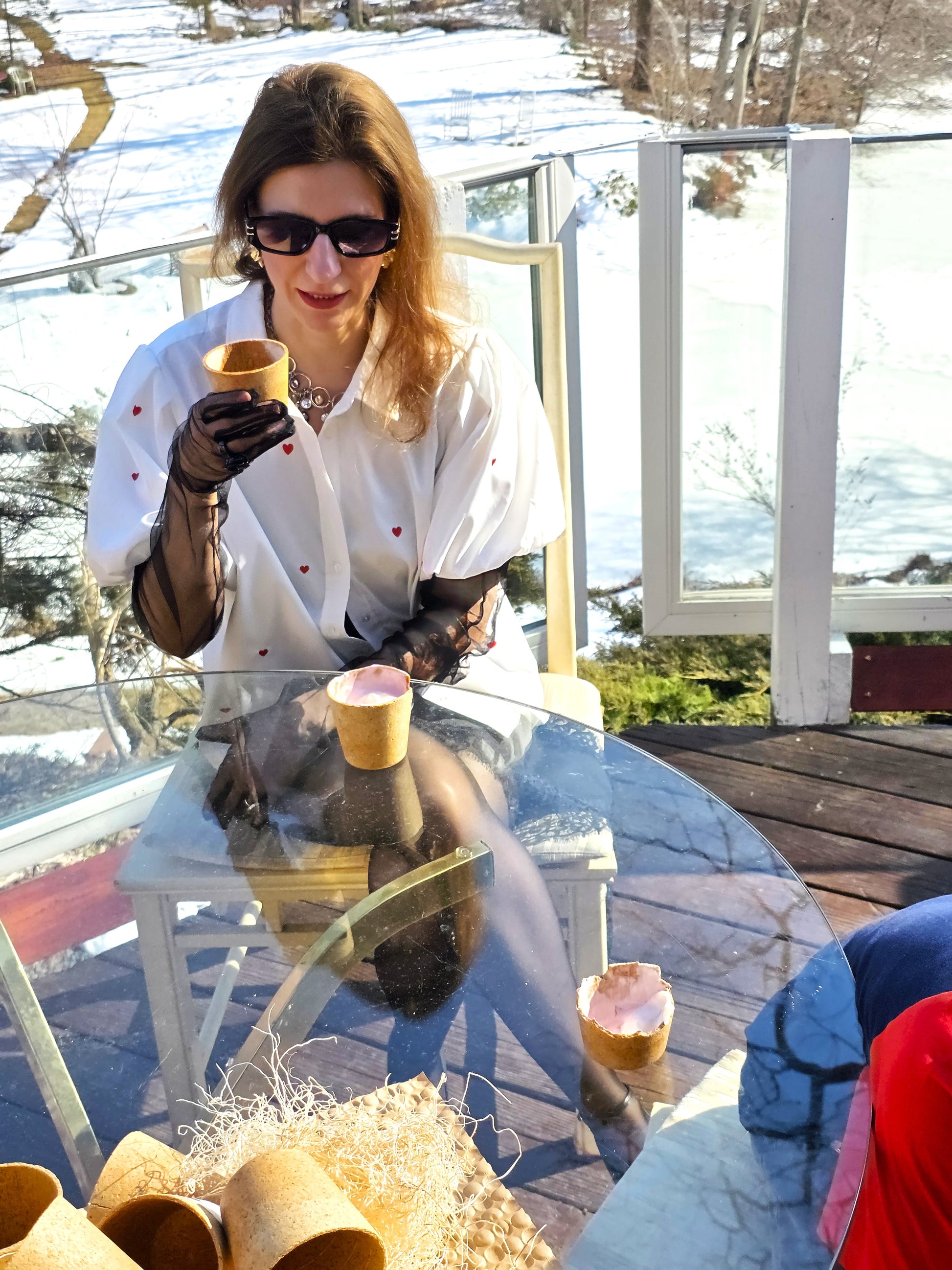 Woman sitting at a glass table outdoors, holding a cup with a snowy landscape in the background.